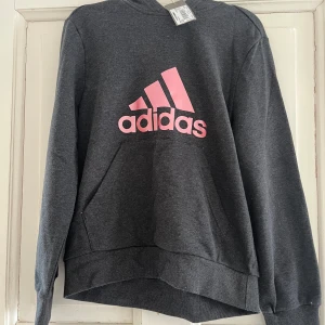 Mörkgrå hoodie från Adidas med rosa logga - Säljer en mörkgrå hoodie från Adidas med stor rosa logga på bröstet. Tröjan har huva, magficka och långa ärmar. Perfekt för en sportig och avslappnad stil. Materialet är mjukt och bekvämt. Oanvänd. Ord pris 479
