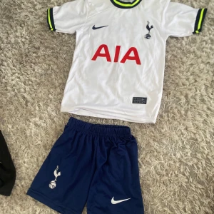 Tottenham Hotspur fotbollströja och shorts - Säljer en vit Tottenham Hotspur fotbollströja med blå Nike-logga, klubbmärke och röd AIA-sponsor på bröstet. Kragen har blå och neongula detaljer med 'SPURS' invävt. Tillhörande blå shorts med klubbmärke och Nike-logga.