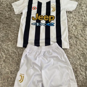 Juventus fotbollströja och shorts - Juventus matchställ med kortärmad tröja i vitt med svarta ränder och guldiga detaljer samt vita shorts med klubbens logga. Tröjan har Jeep och 4XE-tryck på bröstet och rund hals. Materialet är lätt och andas, perfekt för fotbollsträning.
