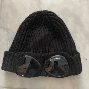 C.P. COMPANY mössa  - Svart stickad mössa från C.P. Company med ribbad struktur och inbyggda goggles framtill. Klassisk modell med uppvikt kant och tydlig logga på glasögonen. Använd flertal gånger men fortfarande i helt okej bra skick! Köpt på NK, taggen går att scanna 💓Nypris var 1200 men säljer för 400 pga att den är väl använd och vill bli av med den👍