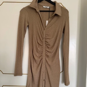 Beige ribbad klänning med knappar från NA-KD - Säljer en beige ribbad klänning från NA-KD med lång ärm och knappar framtill. Klänningen har en snygg rynkad detalj längs knappraden och en klassisk krage. Perfekt för dig som gillar en tight och stilren look.