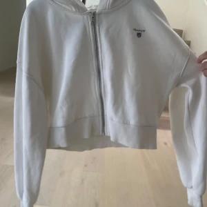 Vit croppad hoodie från Gant - Säljer en jätte fin vit croppad hoodie från Gant med dragkedja framtill och liten logga på bröstet. Tröjan har långa ärmar, huva och en avslappnad passform. Perfekt för en chill och stilren look. Pris kan diskuteras!