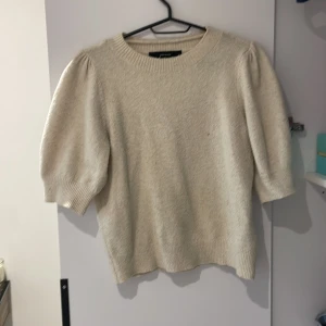 Beige stickad tröja med puffärm från Gina Tricot - Säljer en beige stickad tröja från Gina Tricot med korta puffärmar och rund halsringning. Tröjan har en mjuk och mysig känsla, perfekt för lager-på-lager och passar till både jeans och kjol.