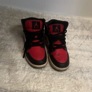 Nike Air Jordans svart/röd/vit - Säljer ett par klassiska Nike Air Jordans i svart, rött och vitt. Skorna har högt skaft, svart snörning och den ikoniska swoosh-loggan i vitt på sidan. Perfekta för dig som gillar streetwear och sneakers med attityd.De är lite sönder där back 