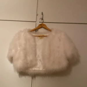 Vit fjäderjacka från Ann Green - Säljer en fluffig och kort vit jacka från Ann Green, tillverkad av fjädrar med polyesterfoder. Jackan har en croppad passform och är perfekt för att sticka ut med sin unika look. Stängs framtill och har korta ärmar.