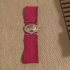 Rosa bälte med silverfärgat spänne - Säljer ett brett rosa bälte med dekorativa kanter och ett ovalt, silverfärgat spänne. Bältet har en elastisk känsla och passar perfekt för att ge outfiten en färgklick. Snygg detalj till klänning eller tunika.