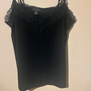 Svart linnetopp med spets från Amisu - Säljer en svart linnetopp från Amisu med tunna axelband och vacker spetsdetalj längs urringningen. Perfekt att styla lager på lager eller bära som den är. Snygg och enkel att matcha till olika outfits.