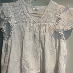 Vit blus med volang  - Säljer en luftig vit blus från Gina Tricot med v-ringning och volangdetaljer på ärmarna. Blusen har korta ärmar och smock upptill, vilket ger en romantisk känsla. Perfekt för varma dagar! Skicka gärna till mig vid vidare frågor😊🫶🩷