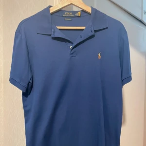 Ralph Lauren pike  - Säljer en klassisk blå pikétröja från Polo Ralph Lauren med broderad logga på bröstet. Tröjan har korta ärmar, krage och knäppning framtill. Modellen är custom slim fit och tyget är i bomull. Nypris ligger på 2000kr. Pris kan diskuteras vid snabb affär. 