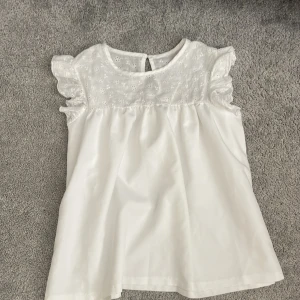 Vit blus med volangärmar - Söt vit blus med blommor upptill och volangärmar. Blusen har en rund halsringning och knapp i nacken. Perfekt för en fräsch och somrig look!