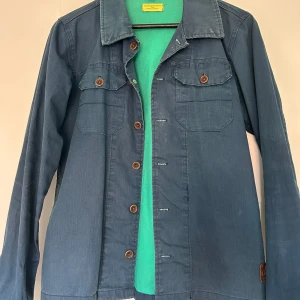 Blå jeansjacka från Scotch & Soda - Säljer en klassisk blå jeansjacka från Scotch & Soda med bröstfickor och knappar framtill. Jackan har en avslappnad passform och är perfekt för lager-på-lager-stil. Snygga detaljer med bruna knappar och krage.