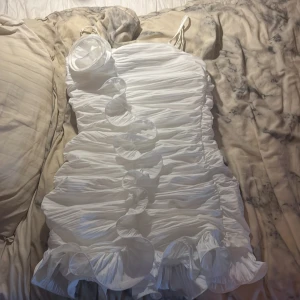 Vit volangklänning från Forever 21 - Säljer en vit kortklänning från Forever 21 med smala axelband och volangdetaljer framtill. Klänningen har en tight passform och är tillverkad i ett lätt, syntetiskt material med rynkade partier för en snygg effekt. Original pris 550 