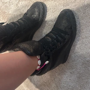 Svarta sneakers från Casadei - Säljer ett par svarta sneakers från Casadei med glittrig yta och snörning. Skorna har en hög modell och platt sula, perfekt för dig som vill sticka ut med en unik look. Snygga detaljer och riktigt cool finish!