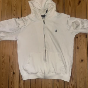 Vit hoodie med dragkedja från Ralph Lauren - Säljer en vit hoodie från Ralph Lauren med dragkedja framtill och klassisk broderad logga på bröstet. Tröjan har huva, långa ärmar och två fickor framtill. Perfekt för en avslappnad och stilren look. Ops är FA-Ke