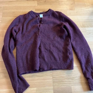 Vinröd kofta från H&M - Mysig vinröd kofta från H&M med rund halsringning och knappar framtill. Tröjan är stickad och har långa ärmar samt ribbade muddar vid hals och ärmslut. Perfekt för lager-på-lager under kyliga dagar.