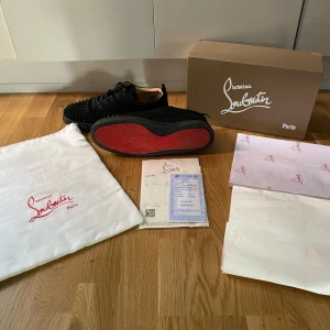 Svarta sneakers med nitar från Christian Louboutin - Säljer ett par svarta sneakers från Christian Louboutin med klassisk röd sula och svarta nitar över tån. Skorna är i mocka och har snörning. Kommer med originalkartong, dustbag och kvitto.