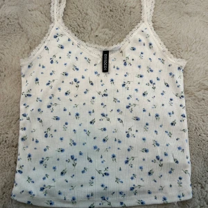 Vit blommig linnetopp med spets från H&M Divided - Supersöt vit linnetopp från H&M Divided med små blå blommor, spetskant och en liten rosett framtill. Perfekt för varma dagar eller som ett lager under en kofta. Tunt ribbat material och smala axelband. Köparen står för frakt och priset går alltid att diskutera🩷