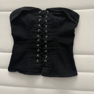 Svart korsett-topp med snörning och dragkedja - Säljer en svart korsett-topp med snörning framtill och dragkedja baktill. Toppen är axelbandslös och har en tight passform som framhäver figuren. Perfekt att styla till olika outfits!