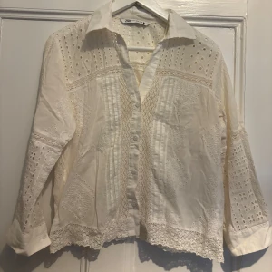 Vit blus med spetsdetaljer från Zara - Säljer en vit blus från Zara med vackra spetsdetaljer och broderade partier. Blusen har lång ärm, klassisk krage och knappar framtill. Perfekt över en bikini på stranden eller cool med ett par jeans.