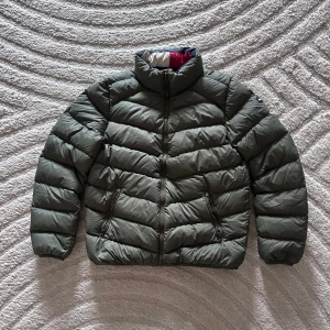 Olivgrön pufferjacka från Tommy Hilfiger - Säljer en olivgrön pufferjacka från Tommy Hilfiger med hög krage och dragkedja framtill. Jackan har långa ärmar och två fickor med dragkedja. Perfekt för kyligare dagar och har en klassisk, sportig look. Jackan har använts ett fåtal gånger och har därför inga hål eller liknande💚