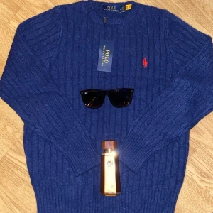 Blå stickad tröja från Polo Ralph Lauren - Säljer en klassisk blå stickad tröja från Polo Ralph Lauren med röd broderad logga på bröstet. Tröjan har rund halsringning och ribbade muddar vid ärmslut och nederkant. Perfekt för en stilren och tidlös look.