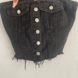 Svart jeans tubtopp med knappar - Cool svart tubtopp i jeansmaterial med silverfärgade knappar framtill och rå kant nertill. Toppen har två fickor med lock och knappdetaljer upptill. Perfekt för en trendig och edgy look.