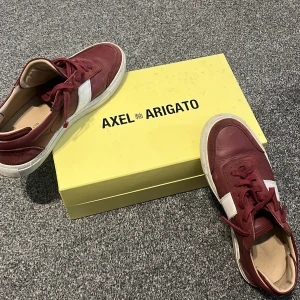 Vinröda sneakers från Axel Arigato - Säljer ett par vinröda sneakers från Axel Arigato med vit sidodetalj och platt sula. Skorna har snörning och kommer med originalkartong och dustbag. Perfekta för dig som gillar stilrena och trendiga sneakers.