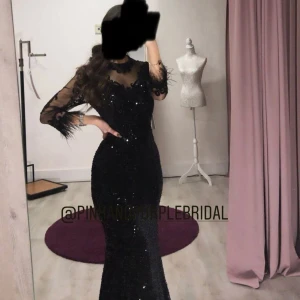 Svart glittrig långklänning med spetsdetaljer - En svart långklänning med glittriga paljetter över hela klänningen. Klänningen har långärmade meshärmar med spets och fjädrar vid ärmslut. Överdelen har dekorativa spetsapplikationer och en elegant, figurnära passform. Perfekt för en glamorös kväll. Oanvänd, köpt i Malmö från Pinkandpurplebridal. Passar som balklänning eller festklänning. 