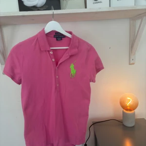 Rosa pikétröja från Ralph Lauren - Säljer en rosa pikétröja från Ralph Lauren med klassisk krage och knappar framtill. Tröjan har korta ärmar och en stor grön broderad logga på bröstet. Perfekt för en sportig och stilren look. Storlek L💕. Priset går att prutas💞