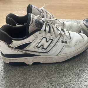 Vita New Balance 550 sneakers - Säljer ett par klassiska New Balance 550 sneakers i vitt med svarta detaljer. Skorna har snörning, rund tå och platt sula. Perfekta för dig som gillar retrostil och streetwear. Ikonisk N-logga på sidan och bekväm passform.