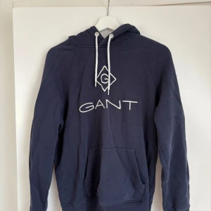 Mörkblå hoodie från GANT - Mörkblå hoodie från GANT med vit broderad logga på bröstet och vita snören i huvan. Klassisk känguruficka framtill och ribbade muddar vid ärmslut och nederkant. Perfekt för en avslappnad stil.