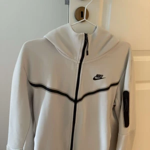 Vit hoodiejacka från Nike med svarta detaljer - Snygg vit hoodiejacka från Nike med svarta detaljer och dragkedja framtill. Jackan har huva, lång ärm och en svart rand som går över bröstet. Perfekt för en sportig och avslappnad stil.