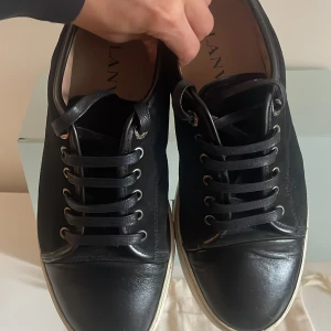 Svarta sneakers från Lanvin - *DISKUTERBART PRIS* Snygga svarta sneakers från Lanvin med vita sulor och klassisk snörning. Skorna har rund tå och är tillverkade i skinn med diskreta detaljer. Perfekta för en stilren och modern look. Storlek 41-42