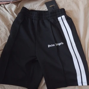 Svarta shorts från Palm Angels med vit rand - Svarta shorts från Palm Angels med vit rand längs sidorna och logga broderad framtill. Elastisk midja och sportig look, perfekta för en avslappnad stil.