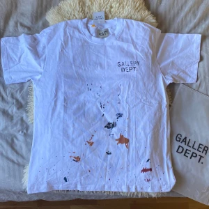 Vit t-shirt från Gallery Dept. med färgstänk - Säljer denna helt nya och enormt snygga vita t-shirt från Gallery Dept. med tryck på bröstet och ryggen. Färgstänk i blått, orange och svart framtill ger en unik look. Riktigt snygg text med skön vibb på ryggen dessutom. Oversized passform och rund hals. Skick 10/10 med tags och bag kvar. Tveka inte på att höra av dig vid frågor eller funderingar! ⭐️