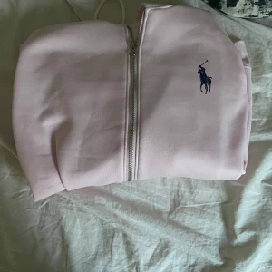 Ljusrosa zip-hoodie från Ralph Lauren - Ljusrosa hoodie med dragkedja från Ralph Lauren. Klassisk design med broderad marinblå logga på bröstet. Perfekt för en avslappnad och stilren look. Skriv vid minsta fundering. Priser kan diskuteras med snabbäffär
