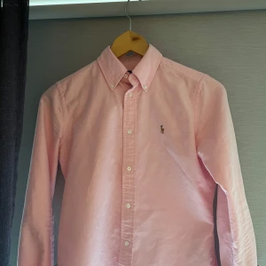 Rosa skjorta från Ralph Lauren - Säljer en ljusrosa skjorta från Ralph Lauren med klassisk button-down krage och broderad logga på bröstet. Passar personer 160-170 ca 😊☀️. Perfekt för dig som gillar stilrena plagg med snygg passform. Vid frågor, tveka inte att fråga 😊👍
