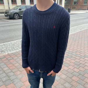 Ralph Lauren cable knit  - Säljer en mörkblå kabelstickad tröja från Ralph Lauren med rund halsringning och diskret röd logga på bröstet. Tröjan har lång ärm och ribbade muddar vid ärmslut och nederkant. Perfekt för lager-på-lager-stil.