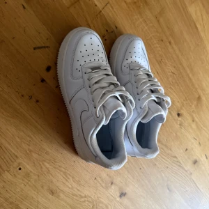 Vita Nike Air Force 1 sneakers - Klassiska vita Nike Air Force 1 sneakers med låg profil och perforerad tå. Skorna har snörning och är tillverkade i skinn med en platt sula. Perfekta för en clean och stilren look.