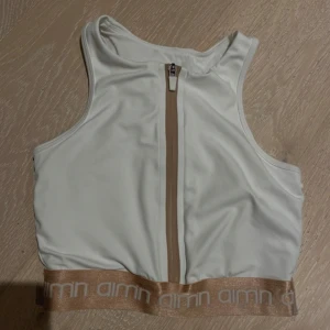 Vit sporttopp Aim'n - Säljer en vit sportig linnetopp från Aim'n med beige resår och logga nertill. Toppen har dragkedja framtill och är ärmlös med rund halsringning. Perfekt för träning eller en aktiv vardag.