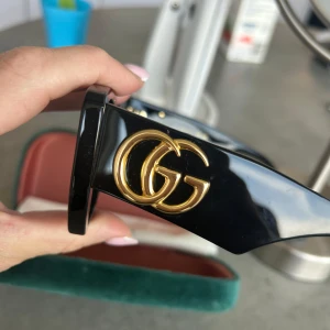 Svarta solglasögon från Gucci nypris 4500 - Stilrena svarta solglasögon från Gucci med breda skalmar och stora guldiga GG-loggor på sidorna. Glasögonen har en fyrkantig form och kommer i originalfodral med putsduk. Perfekta för dig som vill ha en lyxig accessoar.
