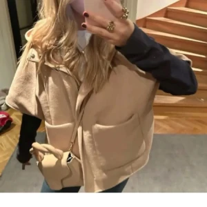 Zara poncho  - Snygg beige capeväst med huva och två stora fickor framtill. Västen har en lös passform och stängs med knappar. Perfekt att slänga över en tröja för en trendig lager-på-lager-look.