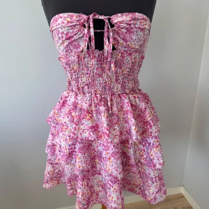 Rosa blommig volangklänning - Söt kort klänning med rosa och vita blommor, volanger och smock i midjan. Axelbandslös med knyt fram och öppen rygg. Den går även att knyta som halterneck. Perfekt för varma dagar och sommarens tillfällen. Storlek M och den är aldrig använd🩷