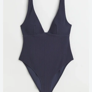 Mörkblå ribbad baddräkt - En stilren mörkblå baddräkt med djup v-ringning och ribbad struktur. Baddräkten har breda axelband och en klassisk passform som ger en tidlös look. Perfekt för strand eller poolhäng.