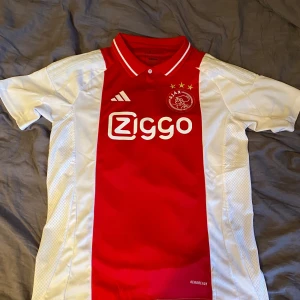 Ajax fotbollströja från Adidas - Säljer en Ajax fotbollströja från Adidas i rött och vitt med klubbmärke och tre stjärnor på bröstet. Tröjan har korta ärmar, klassisk krage och Aeroready-material som andas. Perfekt för dig som älskar fotboll och Ajax!