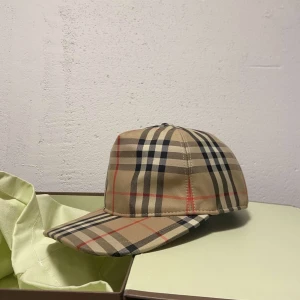 Beige rutig keps från Burberry - Säljer en klassisk beige keps från Burberry med deras ikoniska rutiga mönster i svart, rött och vitt. Kepsen har böjd skärm och justerbar baksida med svart spänne. Perfekt accessoar för en stilren look.