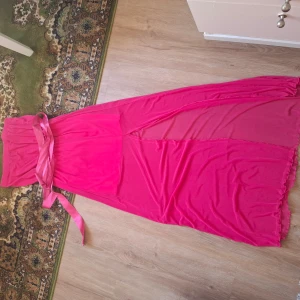 Rosa långklänning med slits och rosett - Säljer en snygg rosa långklänning med markerad midja och bred rosett i satinband. Klänningen har en hög slits på sidan och är tillverkad i ett lätt, flowy material. Perfekt för festliga tillfällen!