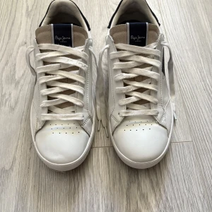 Vita sneakers från Pepe Jeans stl 42 - Snygga vita sneakers från Pepe Jeans med svarta och beige detaljer. Skorna har snörning, rund tå och platt sula. Märkeslogga på sidan och tungan. Perfekta för en stilren och avslappnad look.