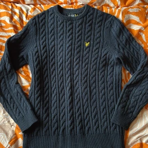 Mörkblå kabelstickad tröja från Lyle & Scott - Säljer en mörkblå kabelstickad tröja från Lyle & Scott med deras klassiska gula logga på bröstet. Tröjan har rund halsringning och långa ärmar, och är sparsamt använd. 
