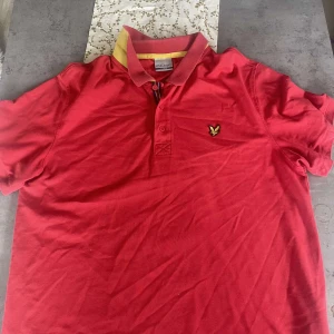 Röd piké från Lyle & Scott - Snygg röd pikétröja från Lyle & Scott med klassisk krage och gul detalj på insidan av kragen. Tröjan har korta ärmar och en broderad logga på bröstet. Perfekt för en stilren och sportig look. Nypris 899 kr, mitt 299 kr